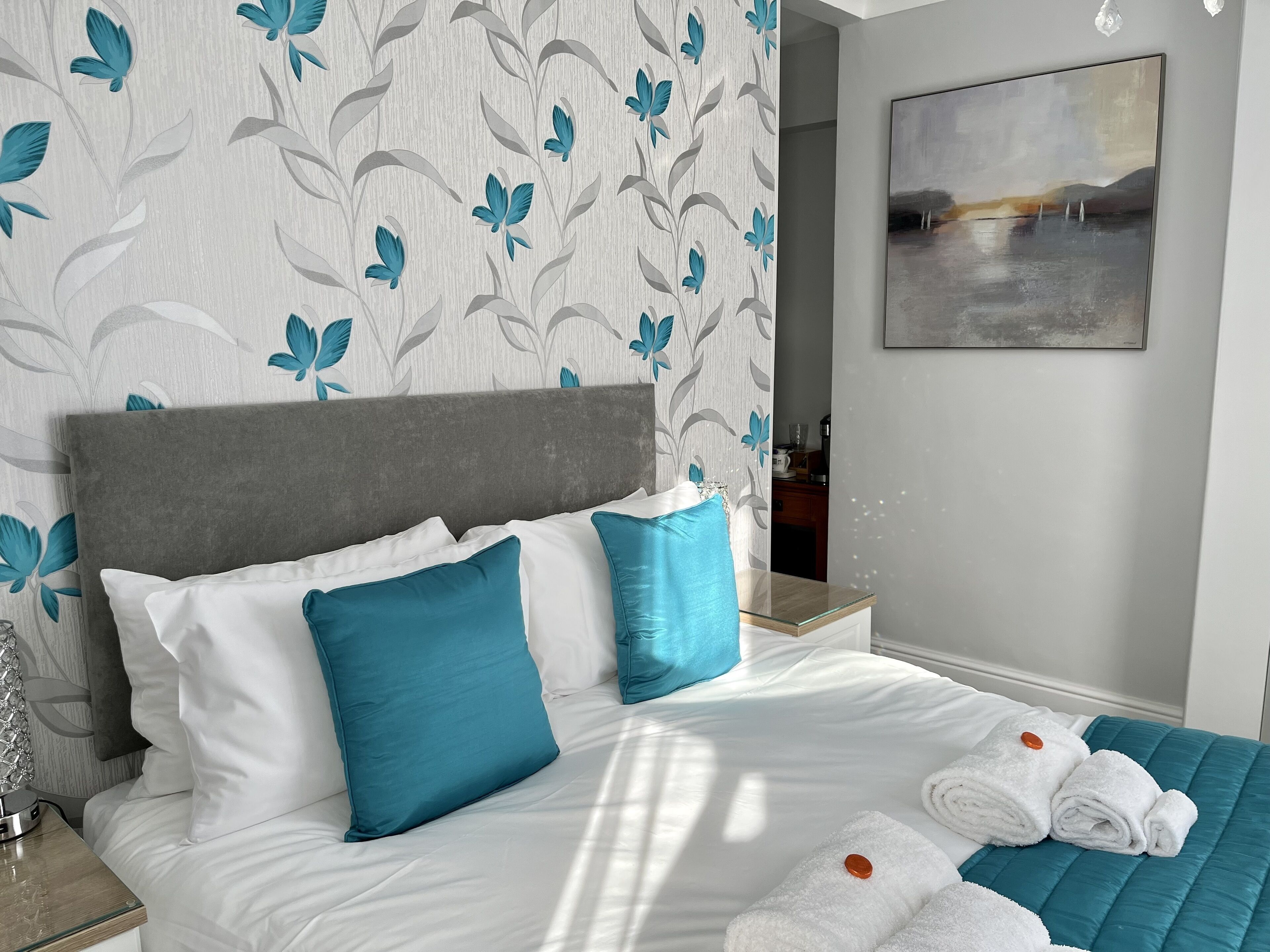 Una cama bien hecha con ropa de cama blanca y azul, un cabecero gris y una almohada decorativa turquesa. Hay un cuadro en la pared y una toalla doblada sobre la cama.
