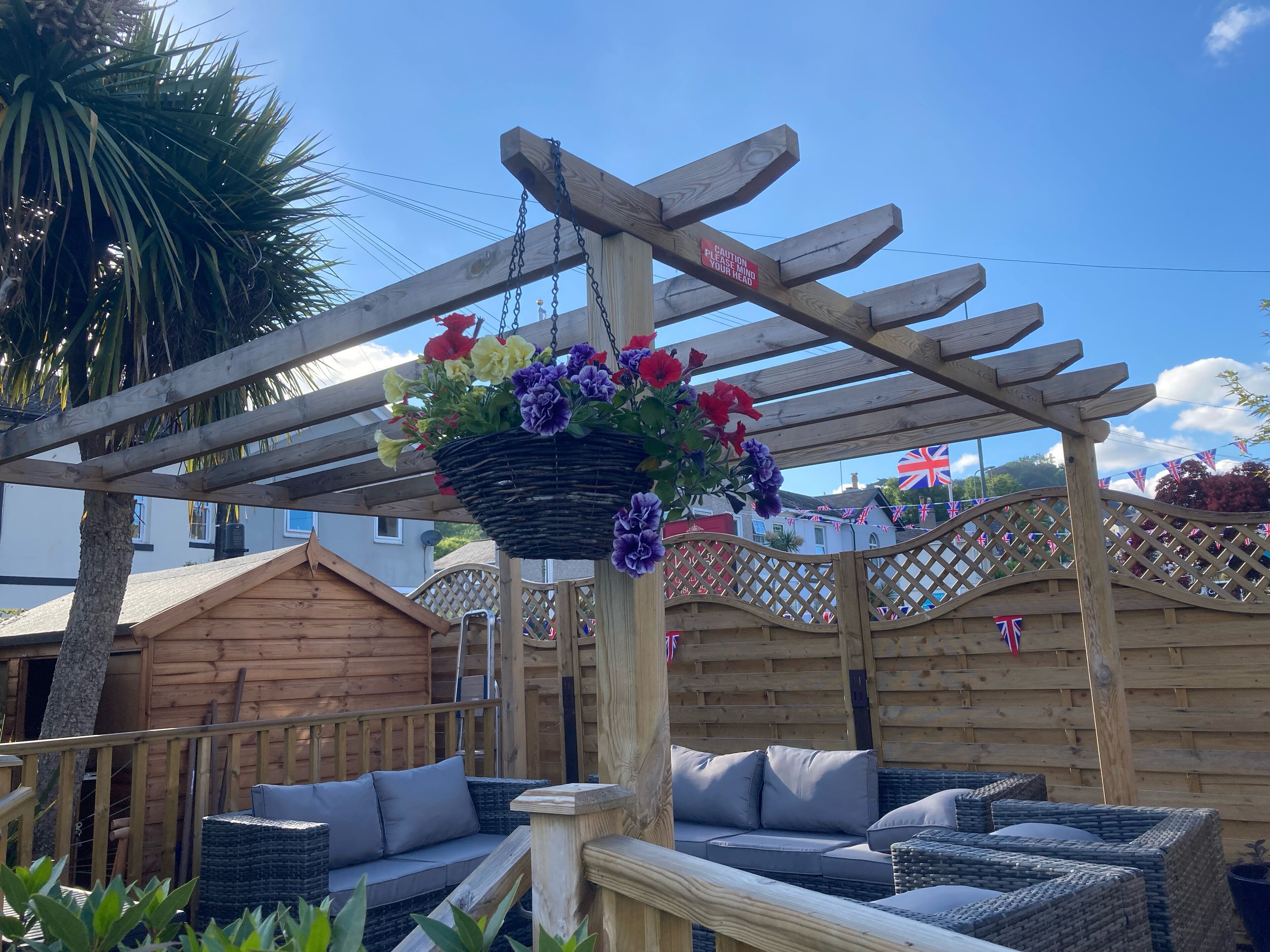A wooden pergola with a hanging basket of flowers, wicker furniture, and a wooden fence with flags.
