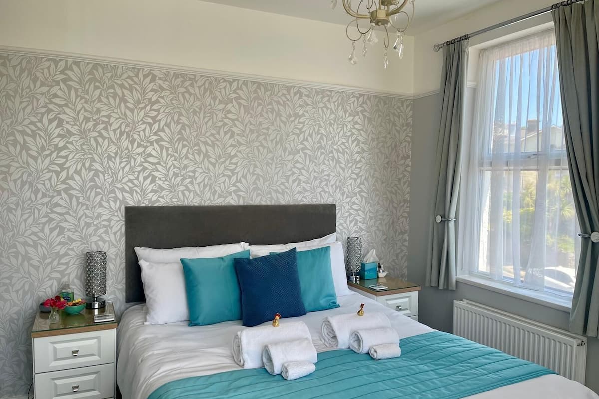 Un lit bien fait, recouvert d’une courtepointe turquoise, avec des oreillers blancs et turquoise, et une tête de lit grise. On aperçoit une table de chevet avec une lampe, un vase de fleurs et une boîte à mouchoirs. Une fen être avec des rideaux gris est visible.