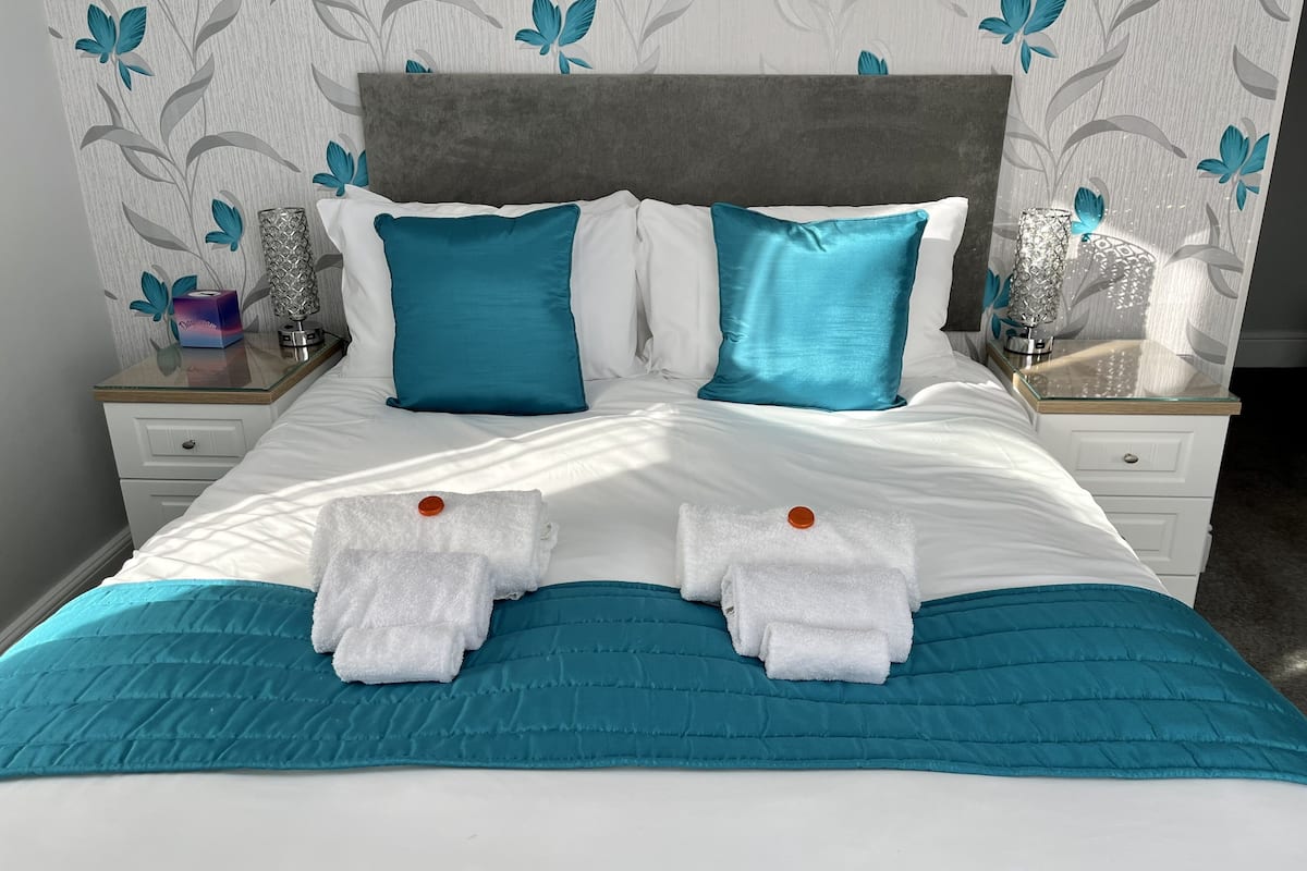 Un lit bien fait, avec une literie turquoise et blanche, deux serviettes et une tête de lit grise.