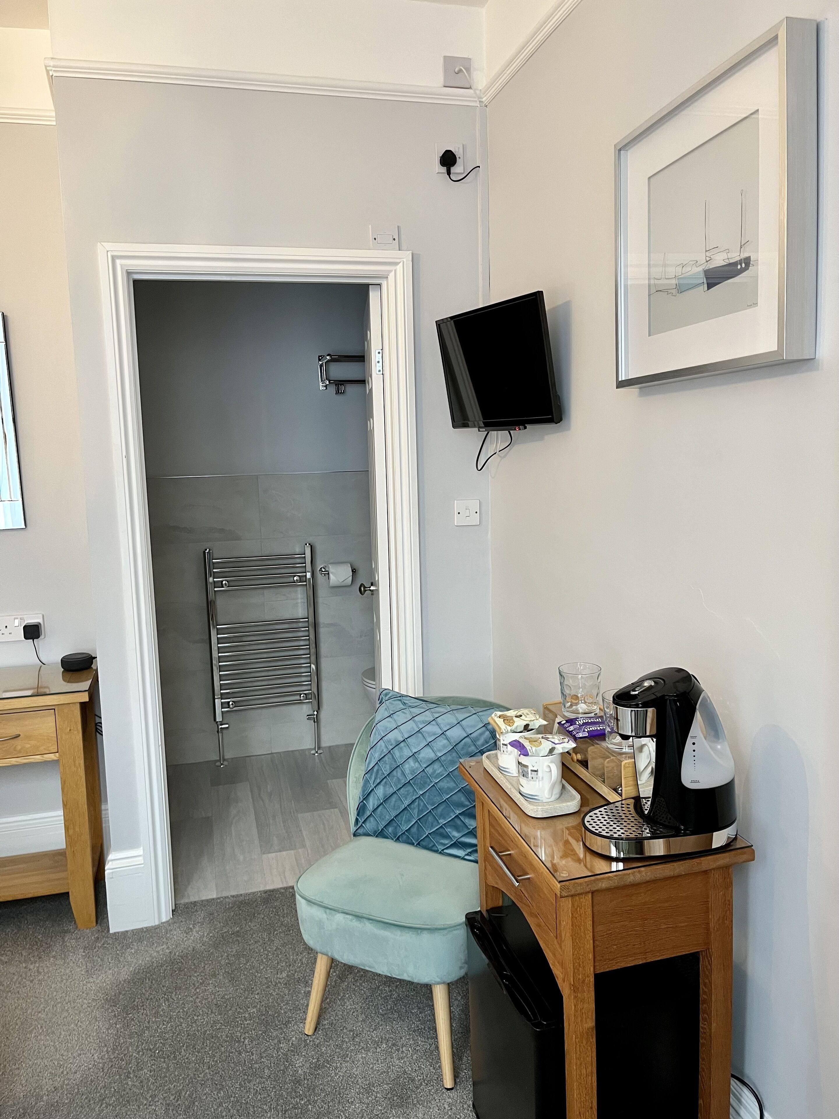 Ein Hotelzimmer mit Schreibtisch aus Holz, einem kleinen Hocker, einer Kaffeemaschine und einem gerahmten Bild an der Wand.