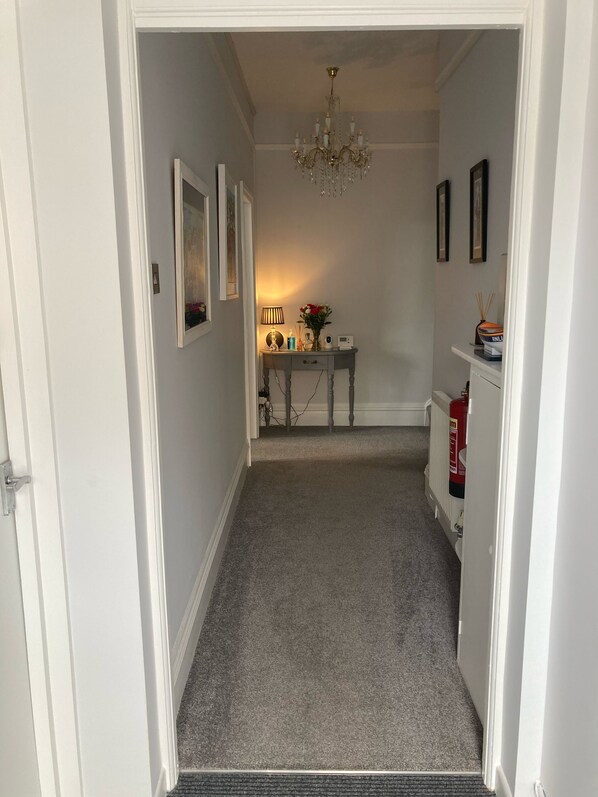 A hallway with a carpeted floor, a chandelier, a table with a lamp, and framed pictures on the wall. - Bentley Lodge Guest House (Torquay)