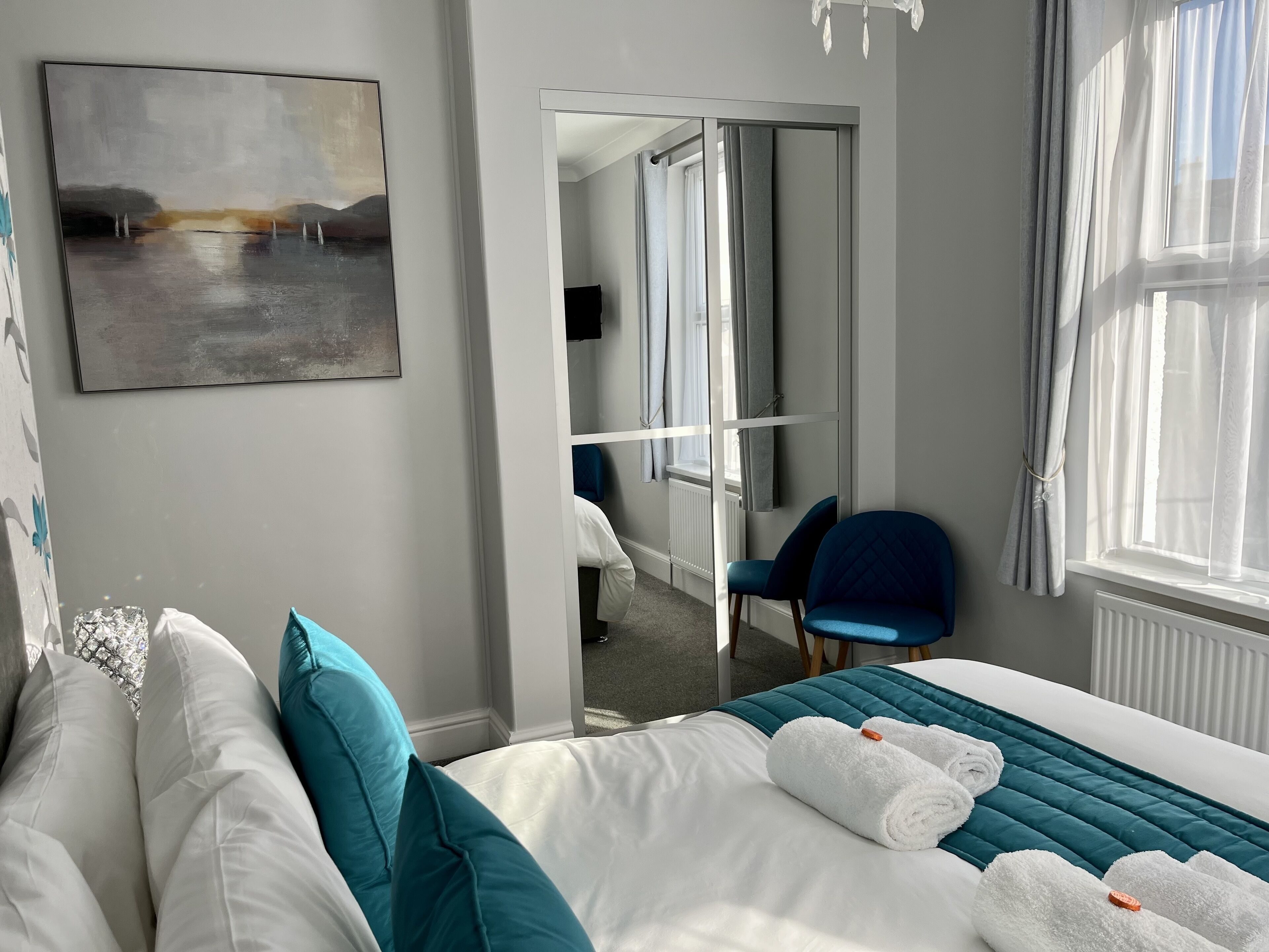 A hotel room with a bed, a chair, a painting, and a view through an open door.