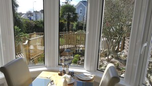 A dining area with a table set for four, large windows, and a view of a garden and neighboring houses. - Bentley Lodge Guest House (Torquay)