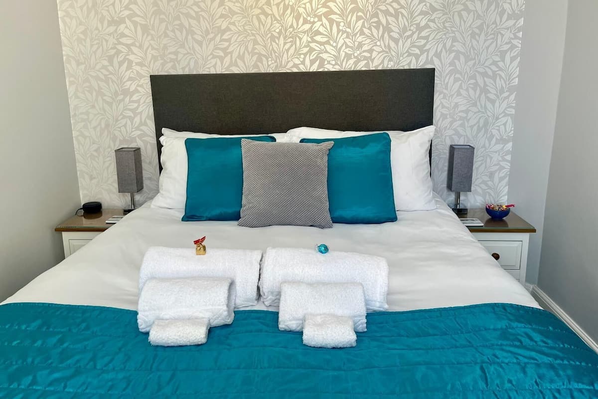 Un lit bien fait, avec une couvre-lit turquoise et des oreillers blancs, une tête de lit et deux tables de chevet ornées de lampes et d’un bol de fruits.