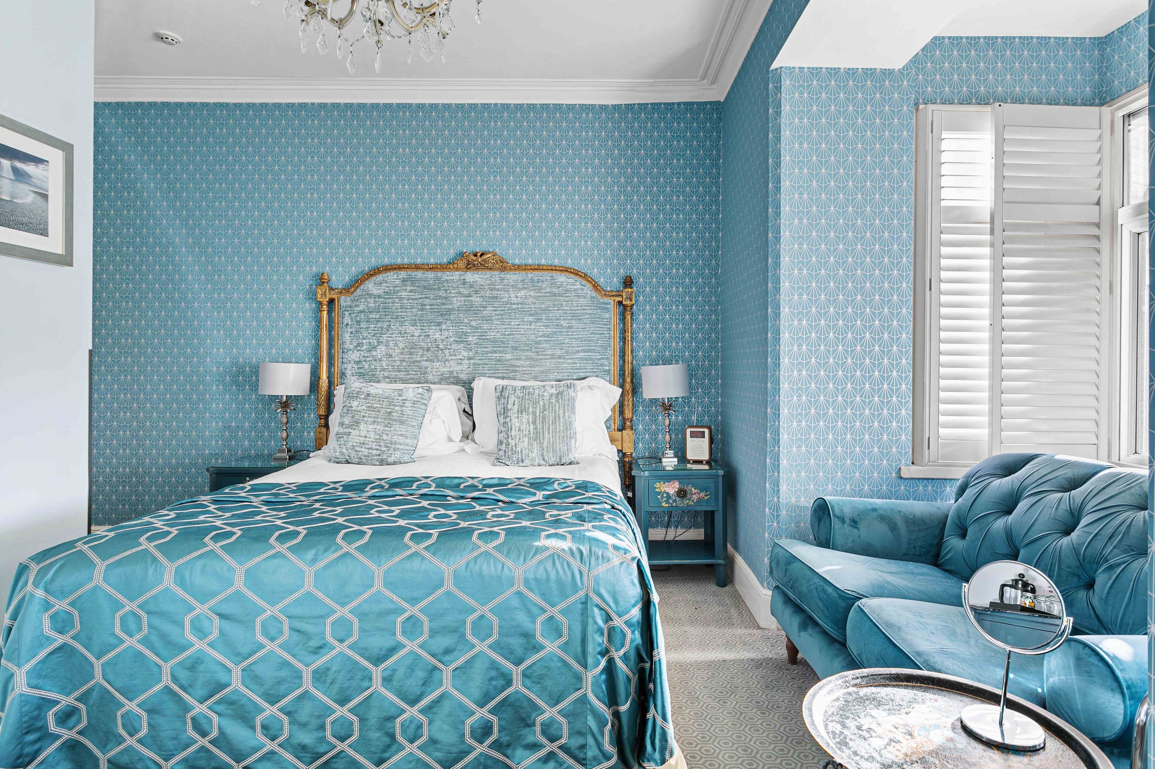 Ein Schlafzimmer mit einem großen Bett, einer Wand mit blauem Muster, einer blauen Couch und einem kleinen, runden Tisch mit Spiegel.