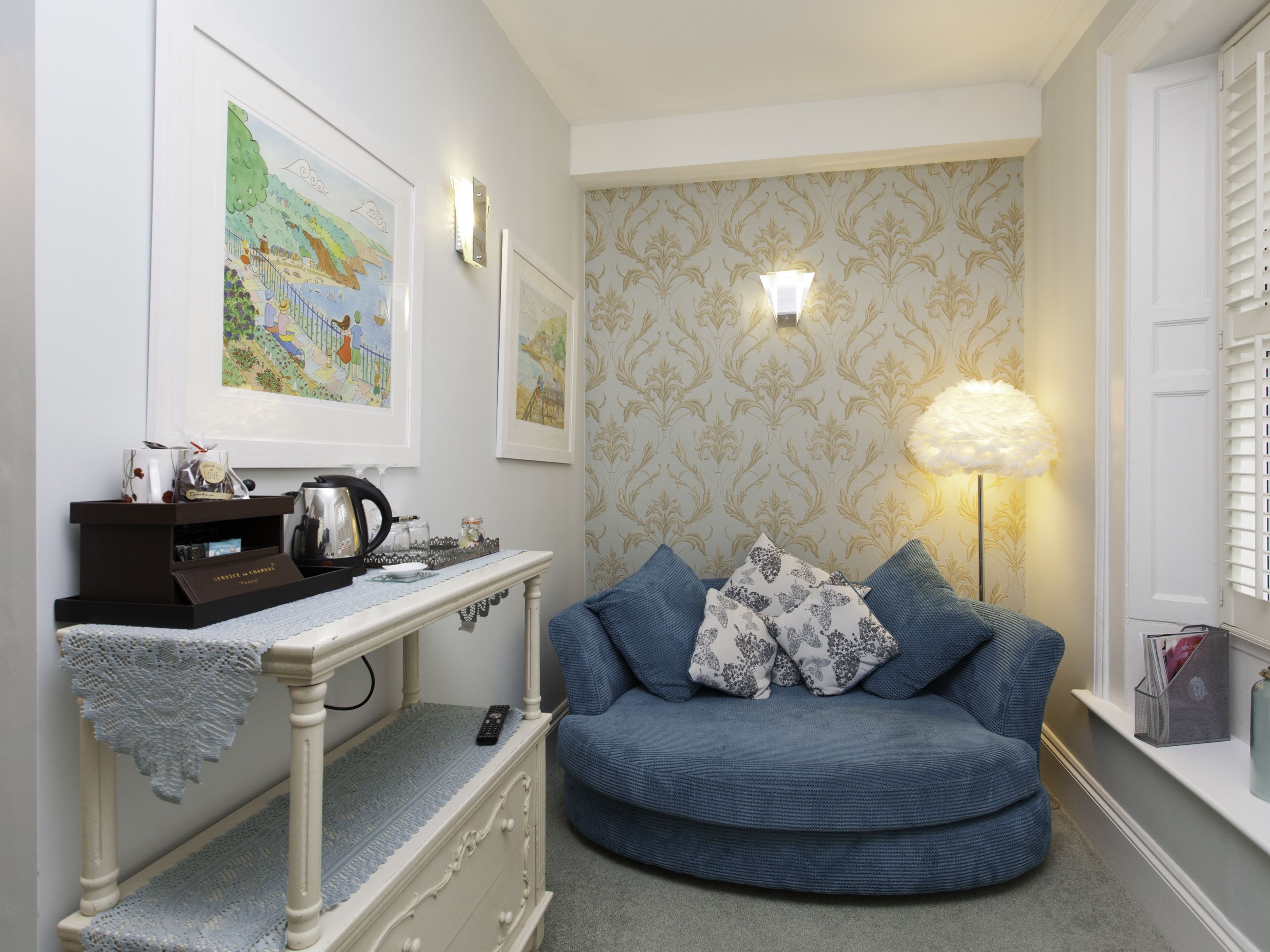 A cozy room with a blue sofa, a small table with a kettle, framed artwork, and a patterned wallpaper.