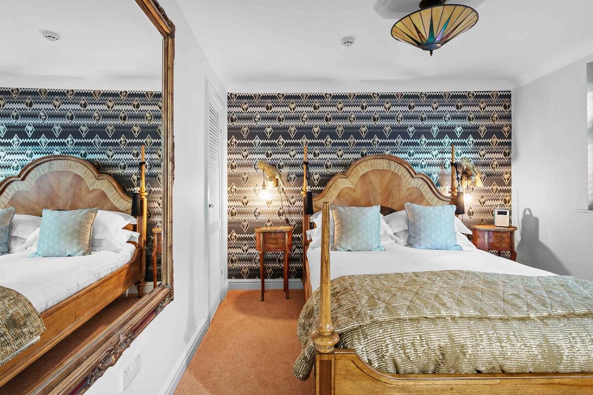 A double bed with a wooden headboard, a bedside table, and a nightstand with a lamp. The room has patterned wallpaper and a ceiling light fixture.