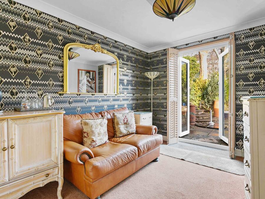 Une pièce avec un papier peint à motifs, un canapé en cuir marron, un grand miroir et une vue sur un espace extérieur, accessible par une porte vitrée.