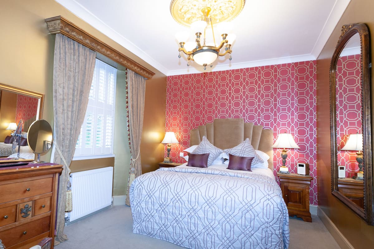 A bedroom with a large bed, two bedside tables with lamps, a dresser, a large mirror, and a window with curtains.