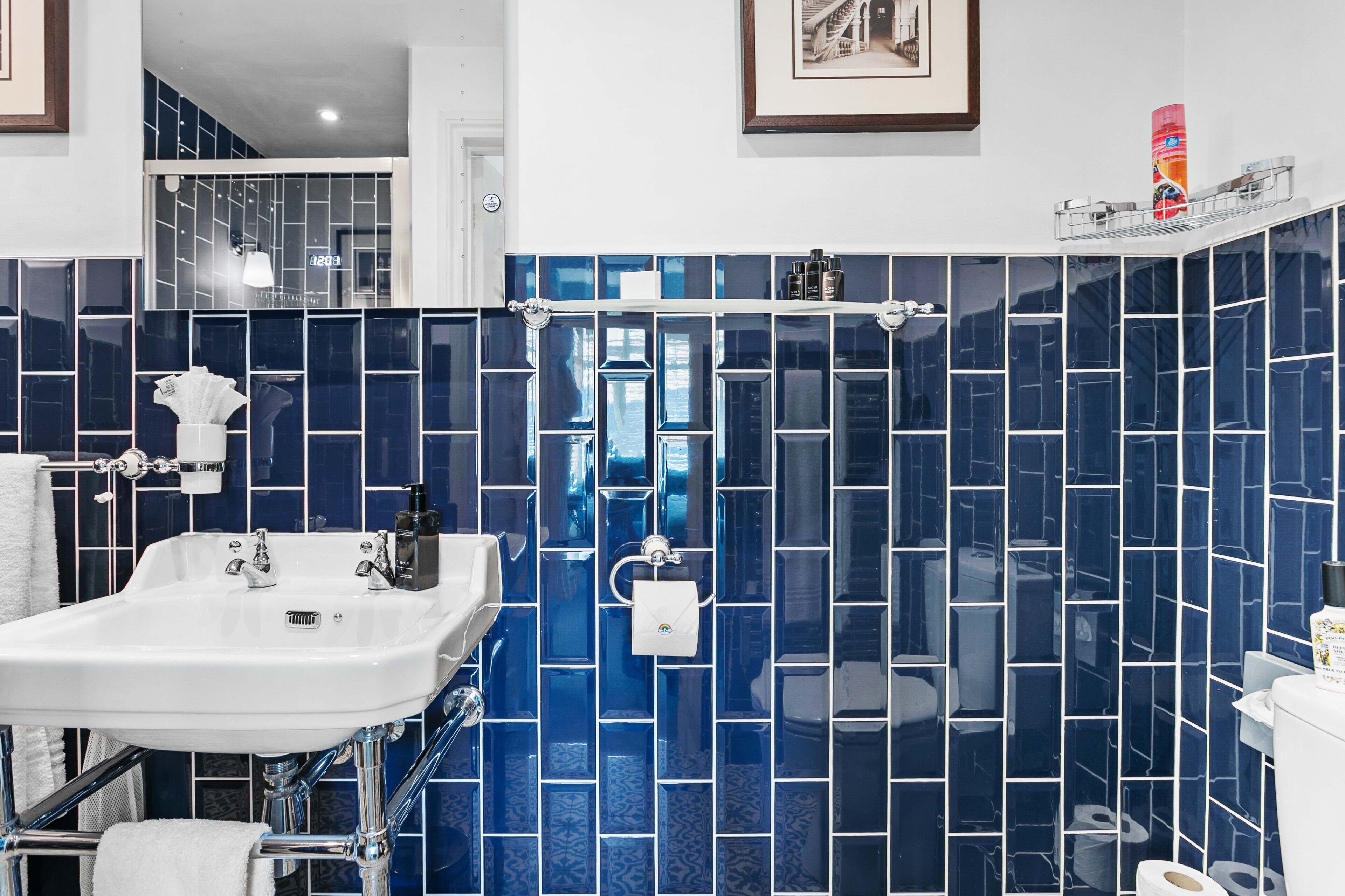 Une salle de bain avec un lavabo blanc, des murs recouverts de carreaux bleus et un tableau encadré au-dessus du miroir.