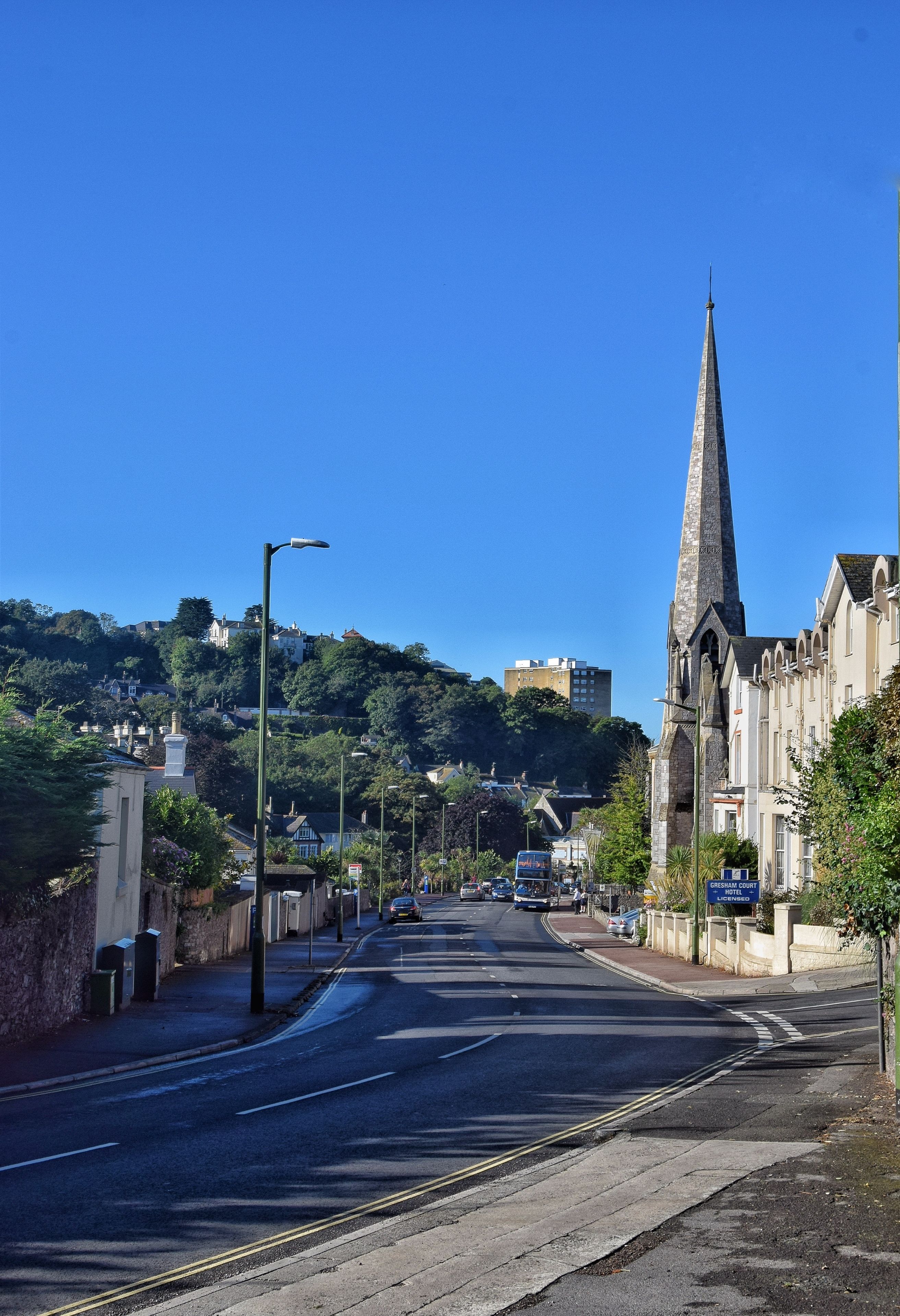 A street with a prominent church spire, a clear blue sky, and a hillside with buildings in the background.