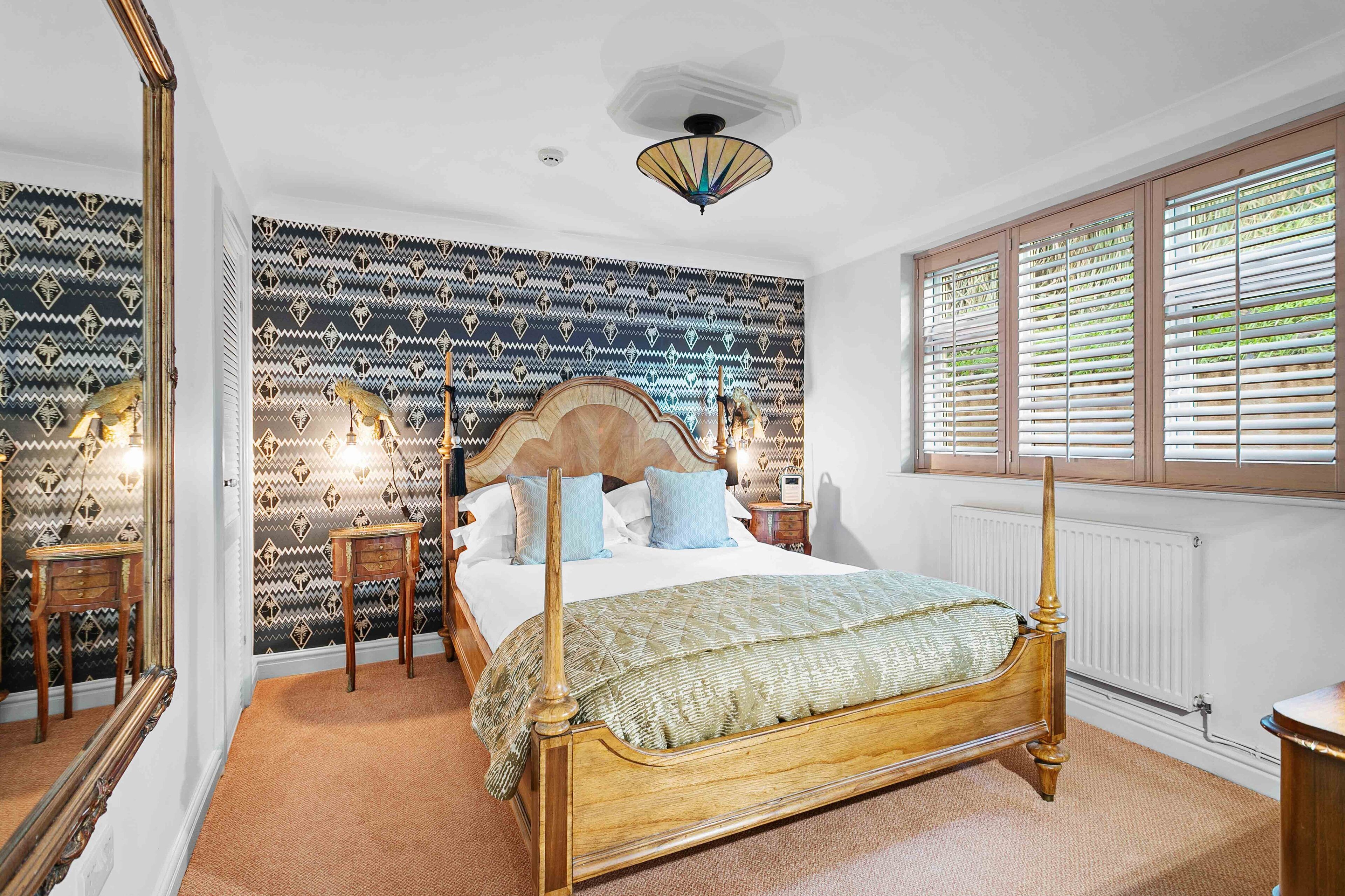 Une chambre à coucher avec un lit à baldaquin, un papier peint à motifs, un ventilateur de plafond et une fenêtre avec des stores.