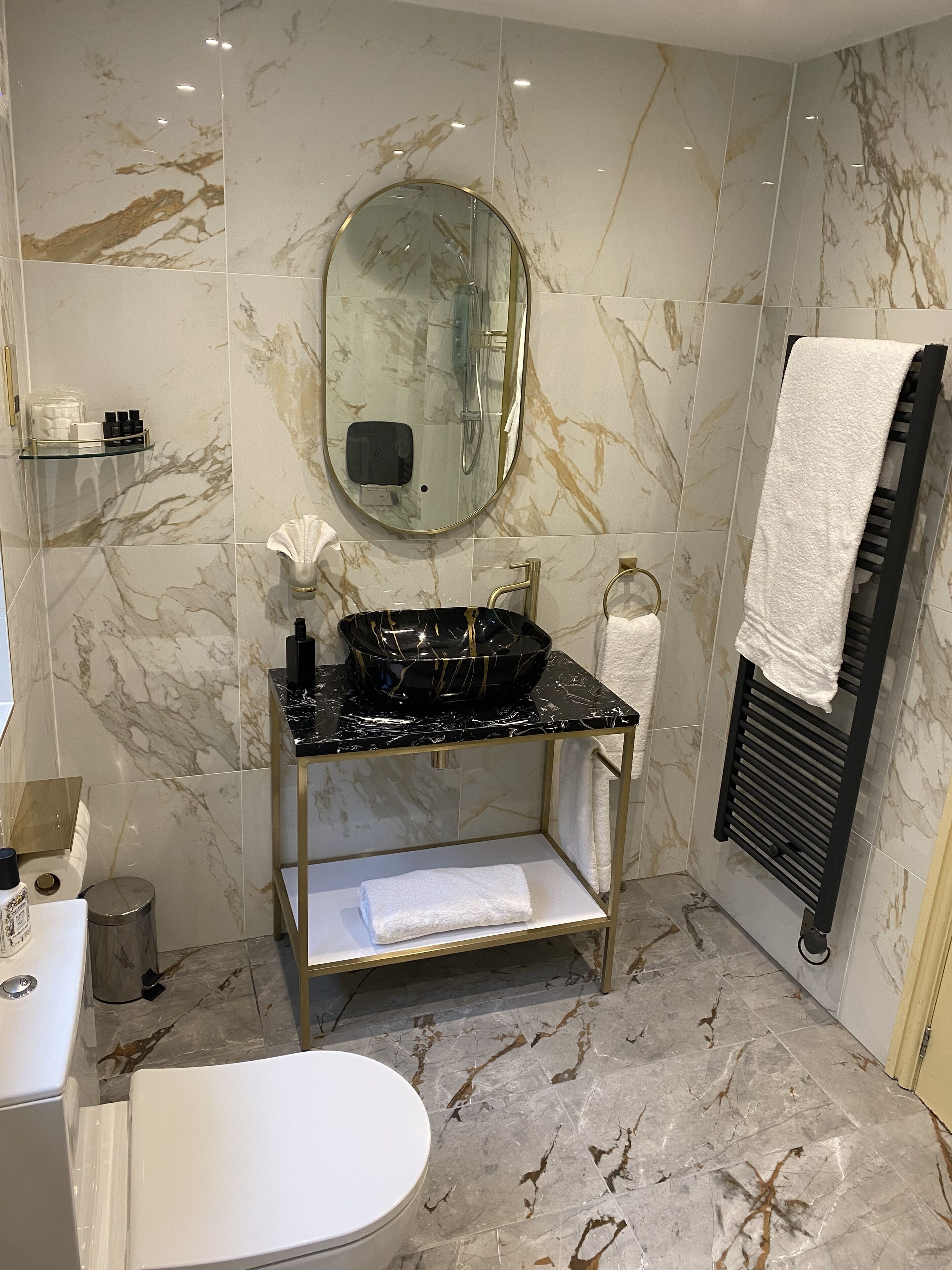 Een badkamer met een marmeren vloer, een ronde spiegel, een zwart wastafel, een handdoekhouder en een toilet.