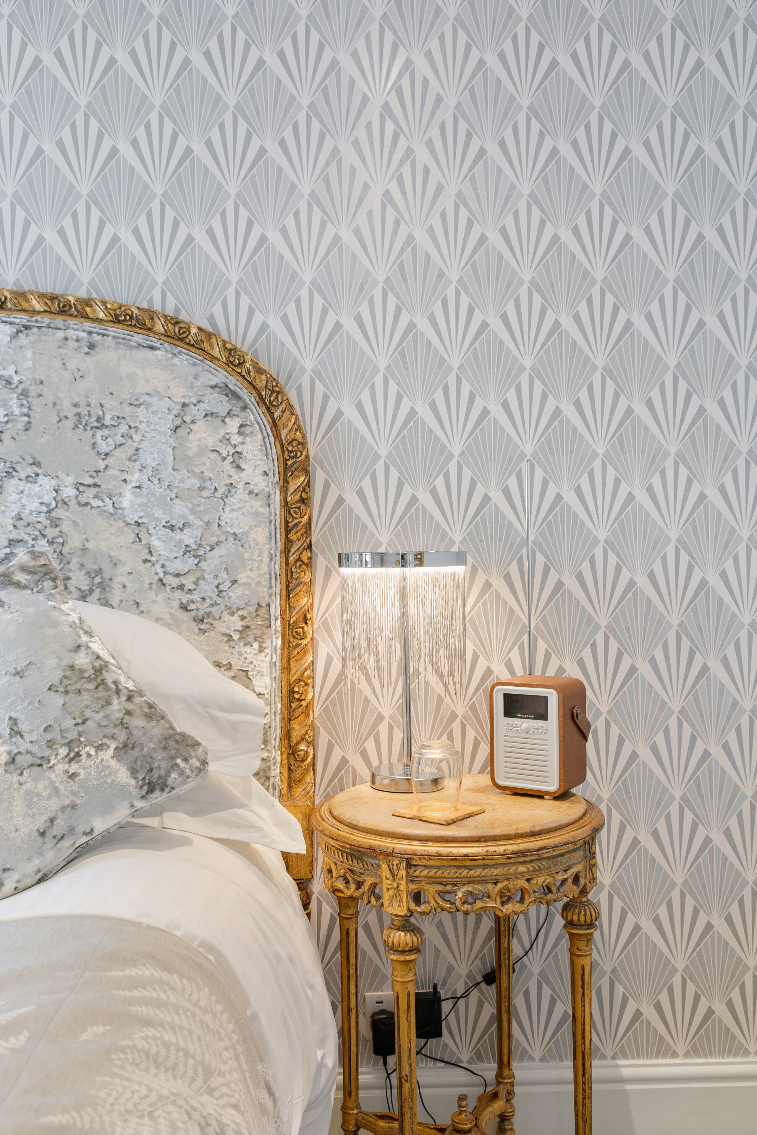 A bed with a marble headboard, a bedside table with a lamp and radio, and a patterned wallpaper background.