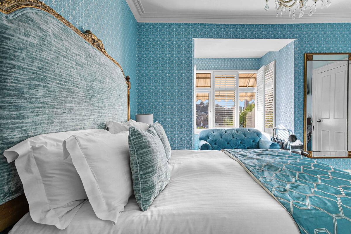 A bedroom with a large bed, a blue patterned wall, and a view of the outside through a window.
