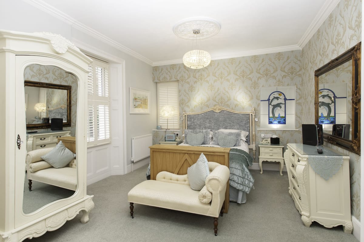 A classic bedroom with a large bed, a vanity with a mirror, a dresser, and a bench.