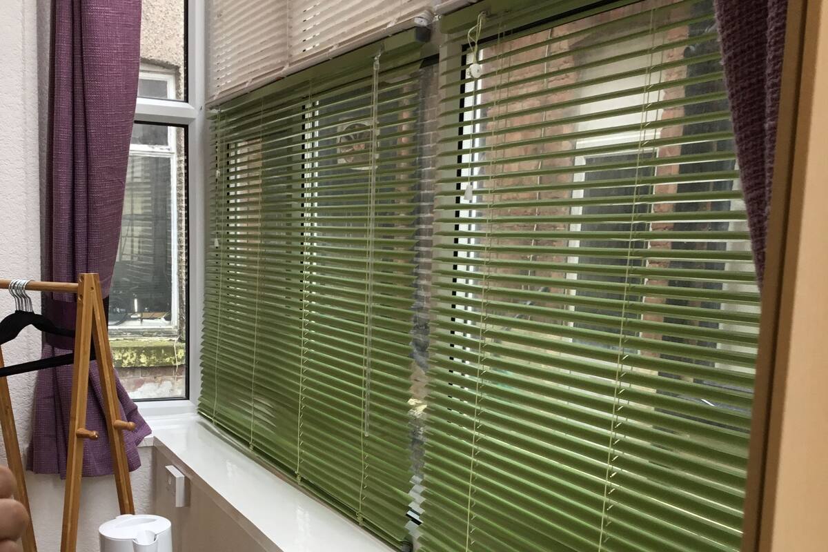 A room with a window covered by green blinds, a small table with a white object, and a purple curtain.