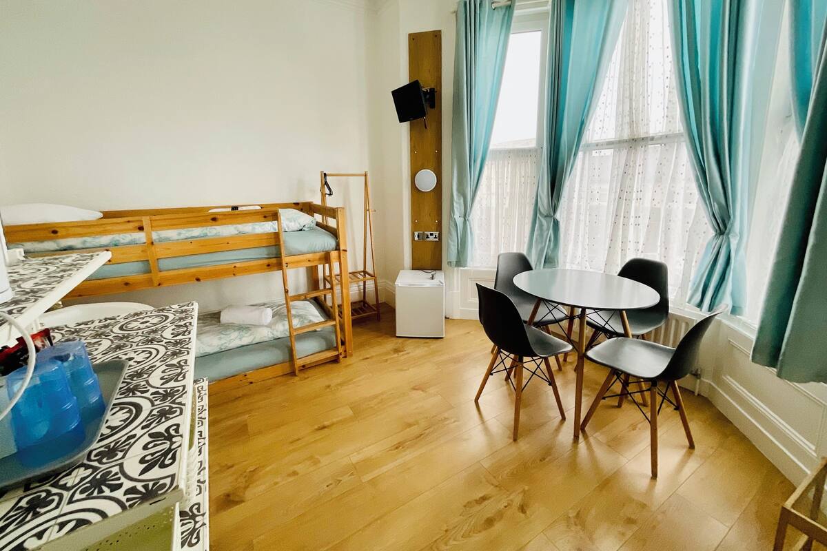 A room with a bunk bed, a small table with chairs, a radiator, and a window with curtains.