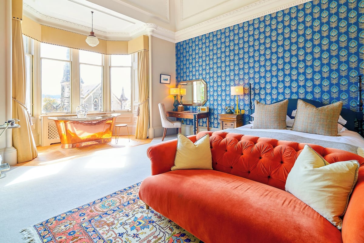 Habitación de hotel con una cama grande, un sofá rojo, un escritorio con una lámpara y vistas a un paisaje urbano a través de amplios ventanales.