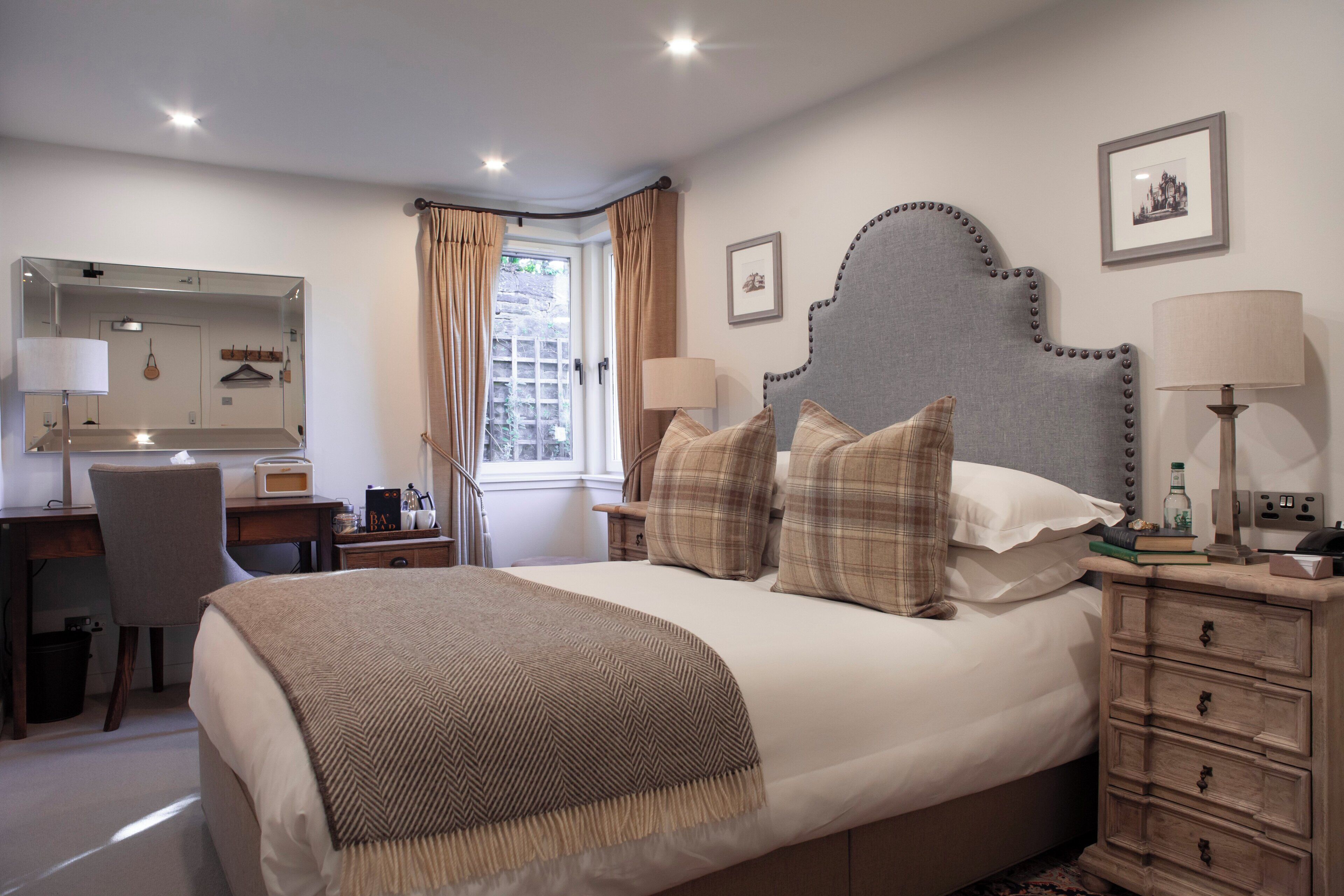A bedroom with a large bed, a desk, a chair, a dresser, a mirror, and a window with curtains.
