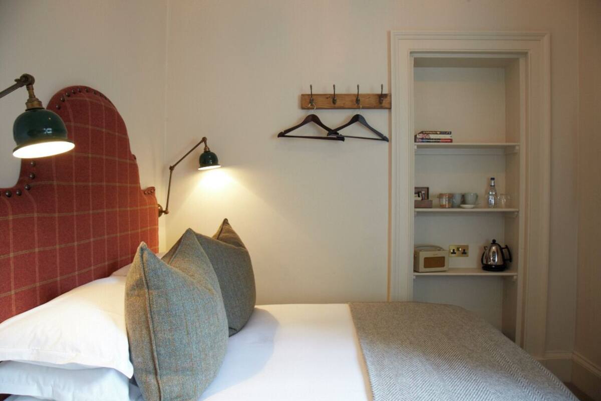 Una cama bien hecha con almohadas, una lámpara de pared y una estantería con libros y un hervidor.