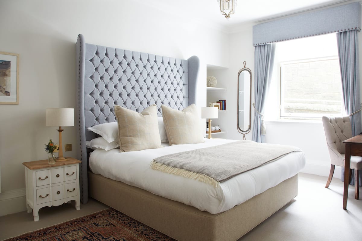 Un dormitorio ordenado con cabecera capitonada, mesita de noche con lámpara, una silla y un amplio ventanal con cortinas.