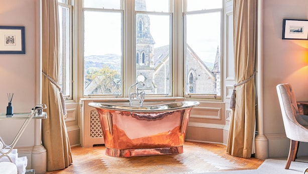 A bathroom with a freestanding copper bathtub, large windows with curtains, and a view of a building outside. - The Roseate Edinburgh (Edinburgh)
