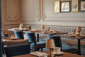 Une salle à manger élégante, avec des tables dressées pour le service, des sièges recouverts d’un tissu bleu et des œuvres d’art encadrées accrochées aux murs.