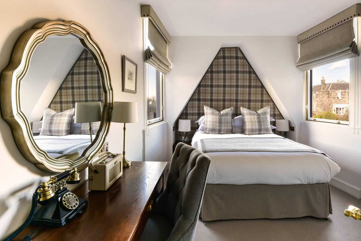 Una habitación de hotel con un espejo grande, un teléfono clásico, un escritorio de madera y una cama con almohadones de cabecera escoceses.