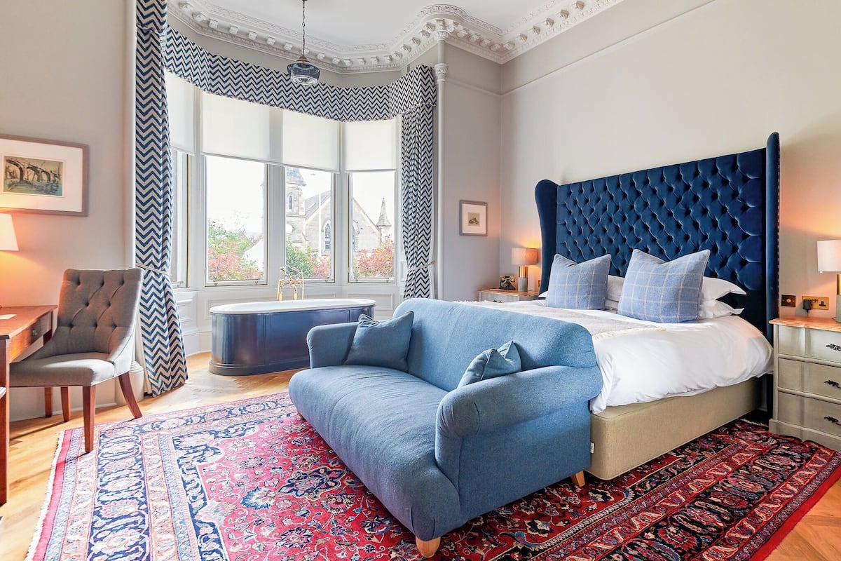Un dormitorio con una cama grande, un sofá tapizado en azul, una silla de madera, un ventanal con vista a un edificio y una alfombra roja estampada.