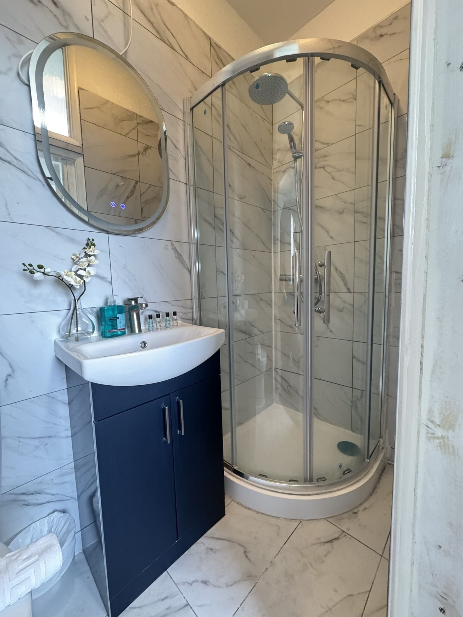 Une salle de bain avec une douche à l’italienne, un lavabo blanc et un meuble sombre, ainsi qu’un miroir rond accroché au mur.
