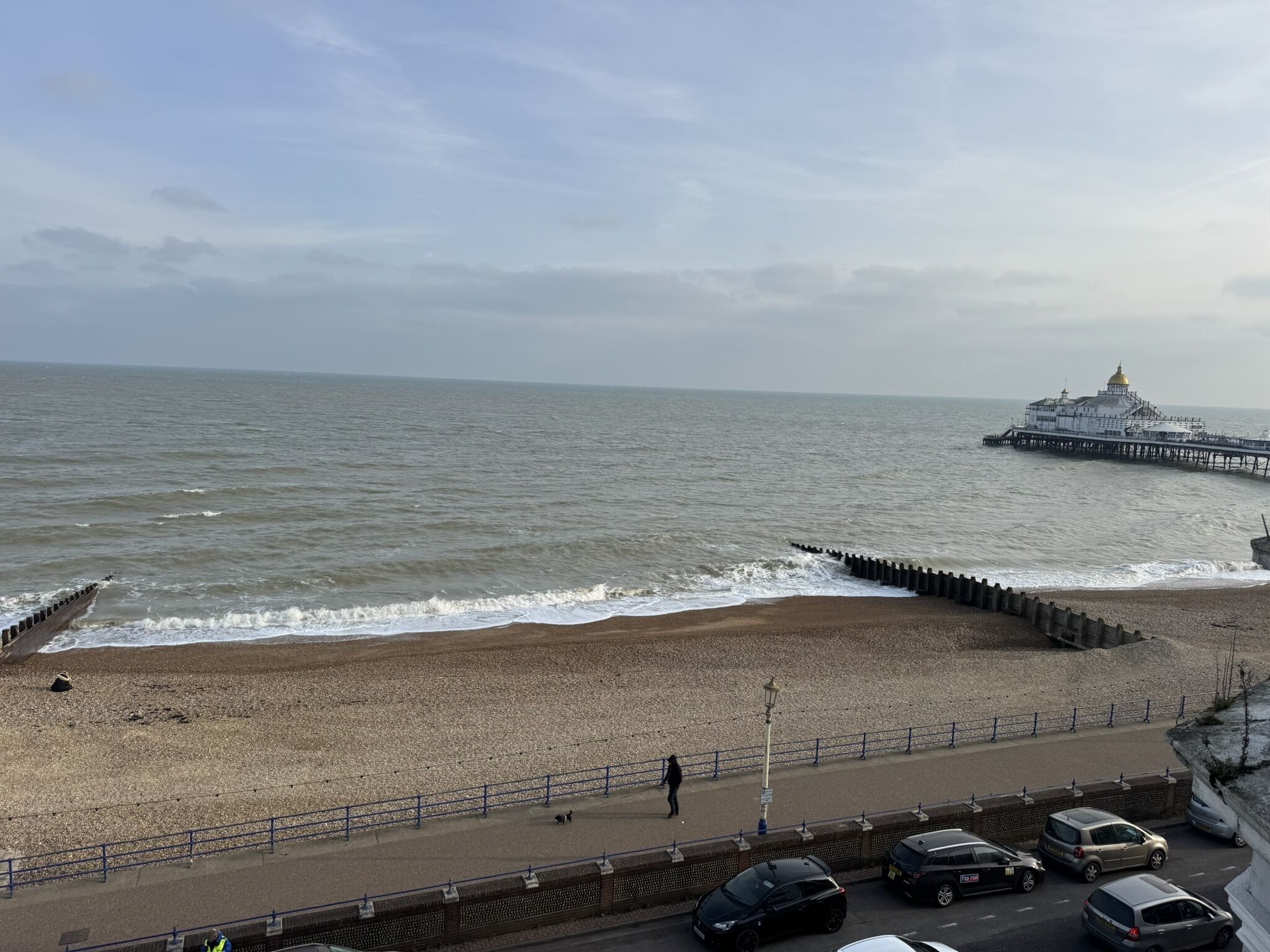 Une plage avec une promenade, une jetée surmontée d’un dôme doré et des voitures garées le long de la route.