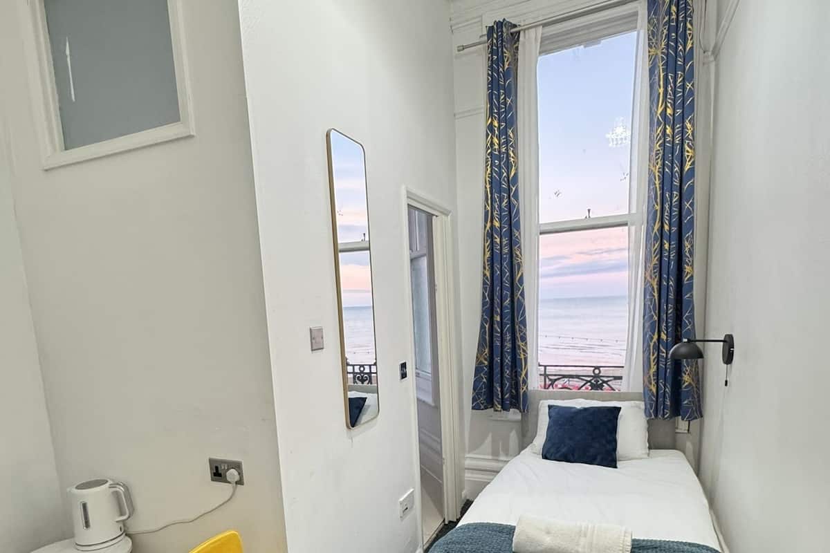 A hotel room with a bed, a small desk, a chair, a window with curtains, and a view of the sea.
