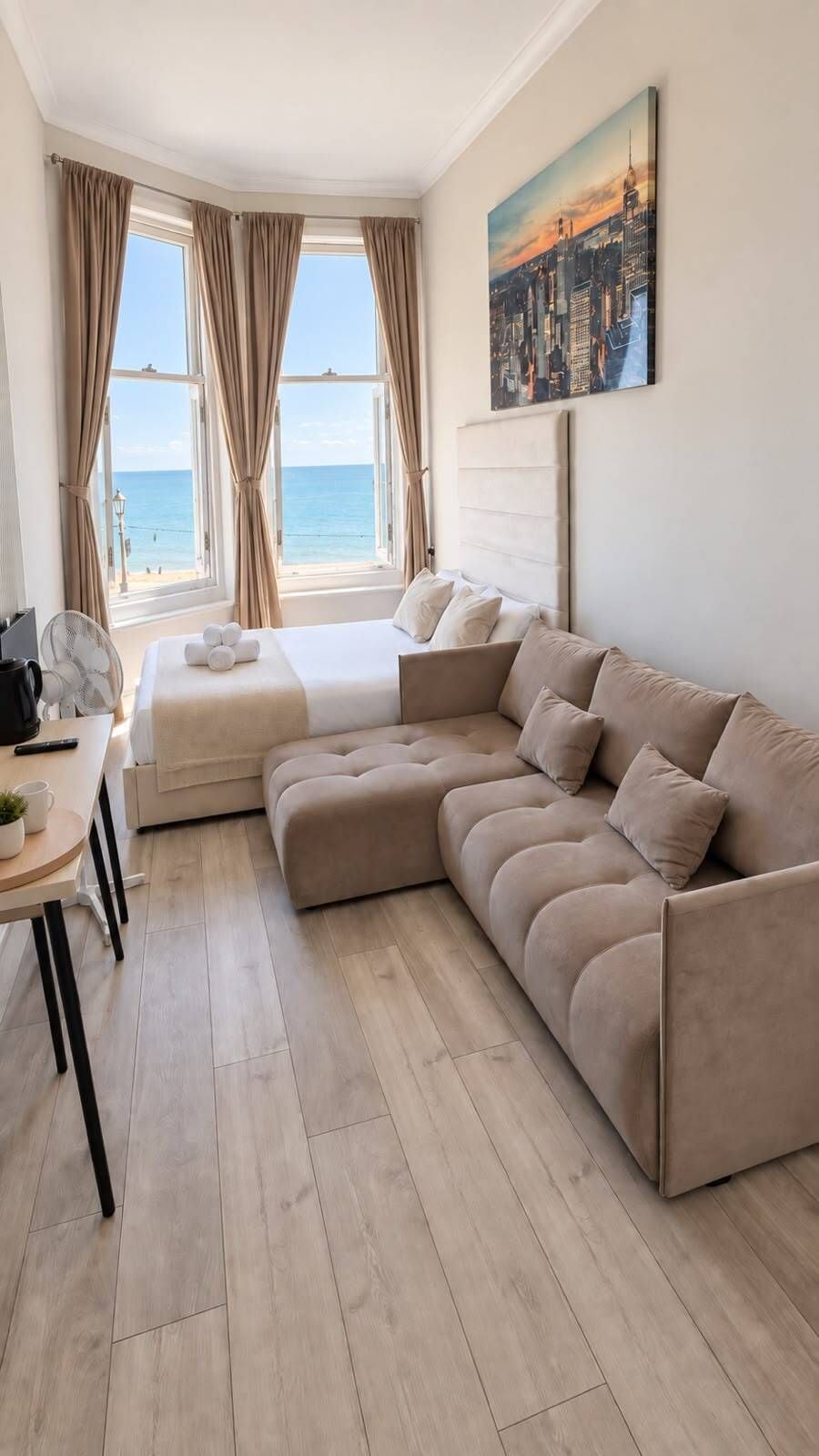 Un dormitorio moderno con una cama grande, un sofá, un escritorio con un ventilador y un cuadro de un paisaje urbano.