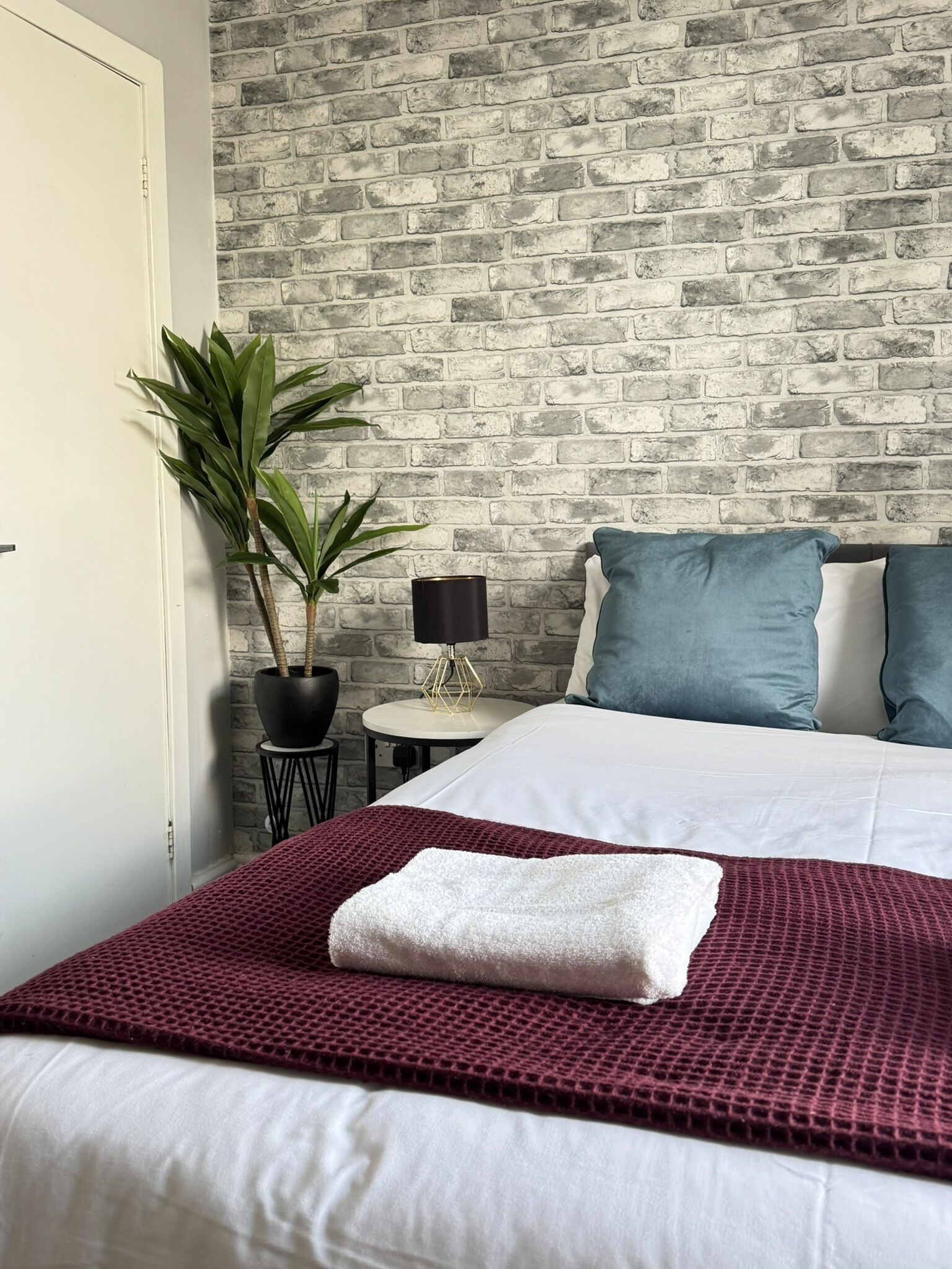A neatly made bed with a white and red blanket, a potted plant, a small round table with a lamp, and a brick-patterned wall.