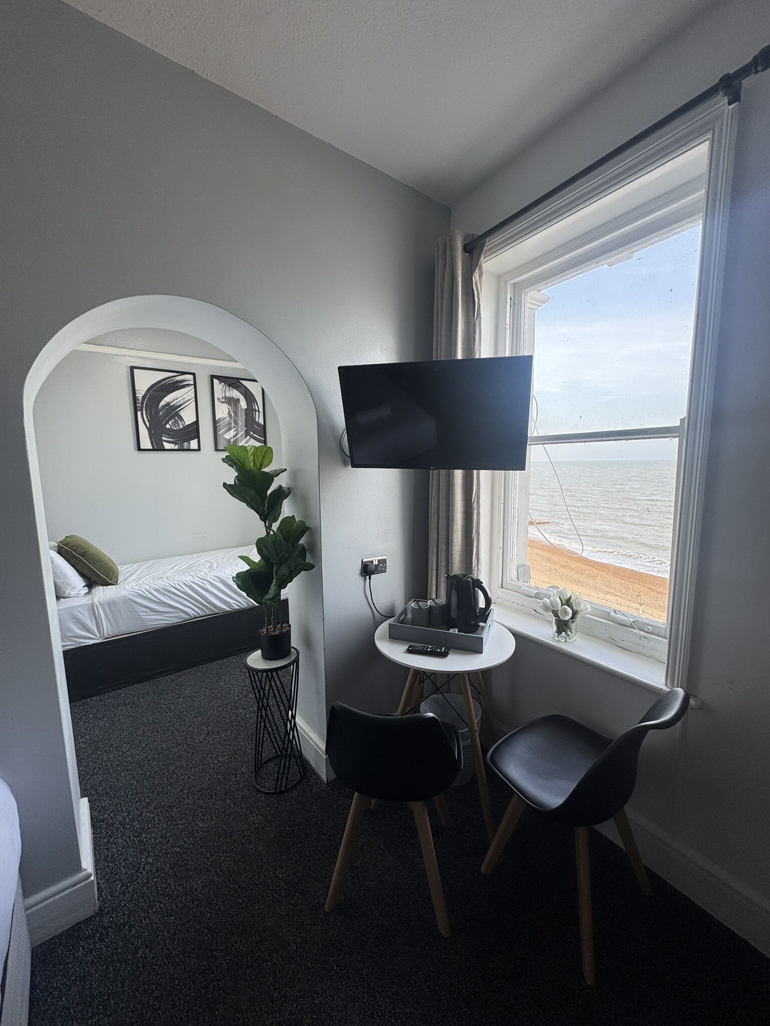 A hotel room with a bed, a TV, a desk with a chair, a potted plant, and a window with a beach view.