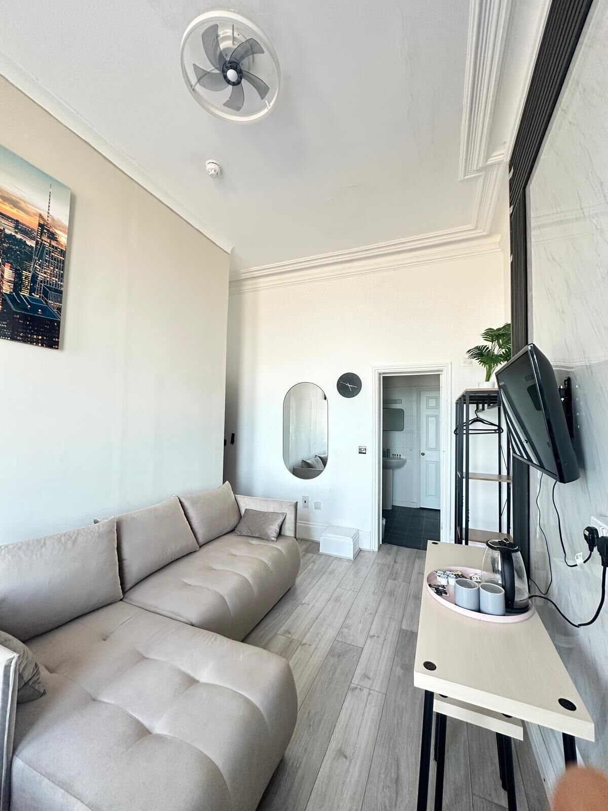 A modern living room with a beige sofa, a standing fan, a flat-screen TV mounted on the wall, and a cityscape painting on the wall.