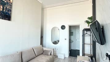 A modern living room with a beige sofa, a standing fan, a flat-screen TV mounted on the wall, and a cityscape painting on the wall.
