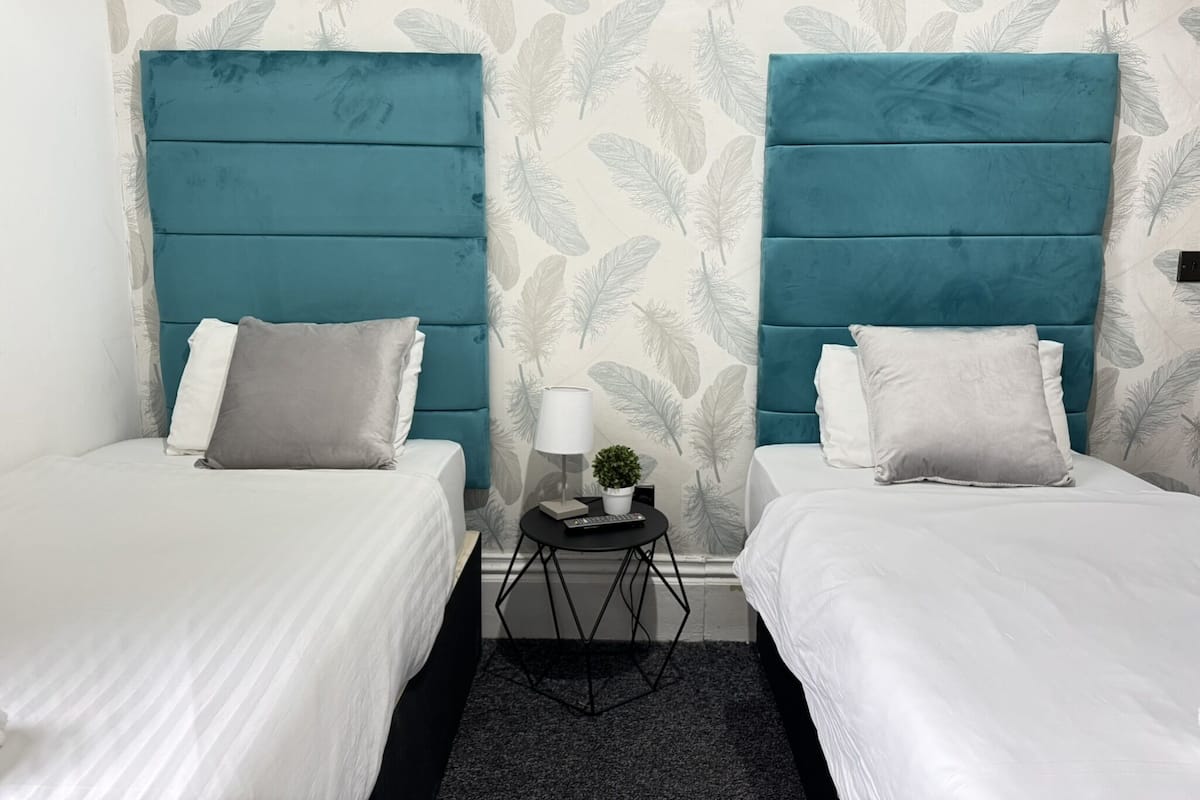 A hotel room with two beds, each with a teal headboard, white bedding, and grey pillows. A small round table with a lamp and a plant is between the beds.