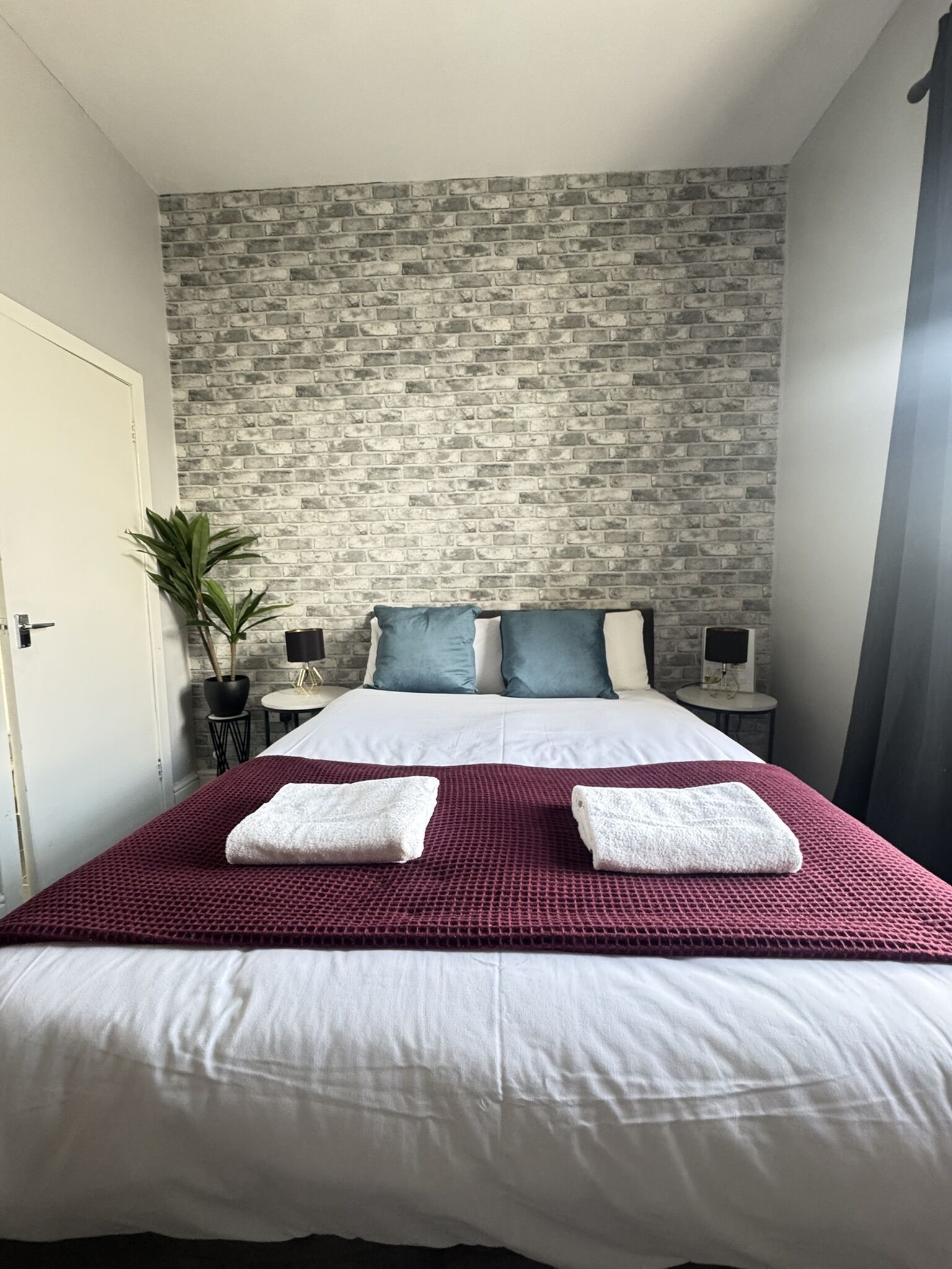 A hotel room with a brick-patterned wall, a large bed with white linens, two bedside tables with lamps, and a potted plant.