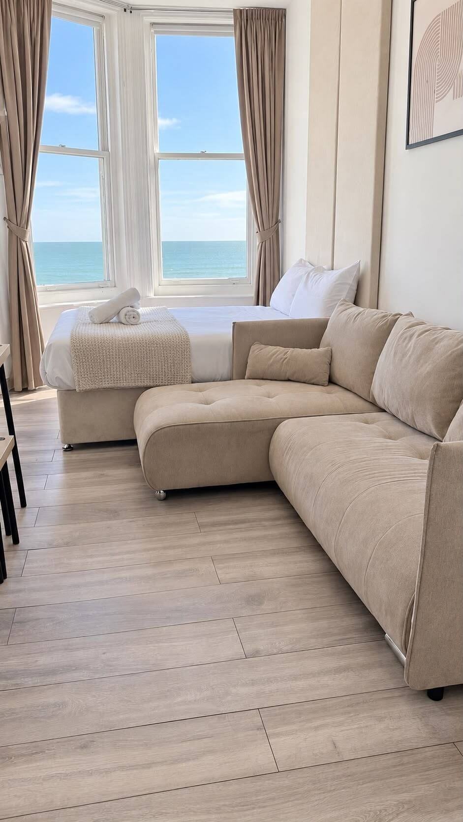 A modern living room with a beige sectional sofa, a small ottoman, and a view of the ocean through large windows.