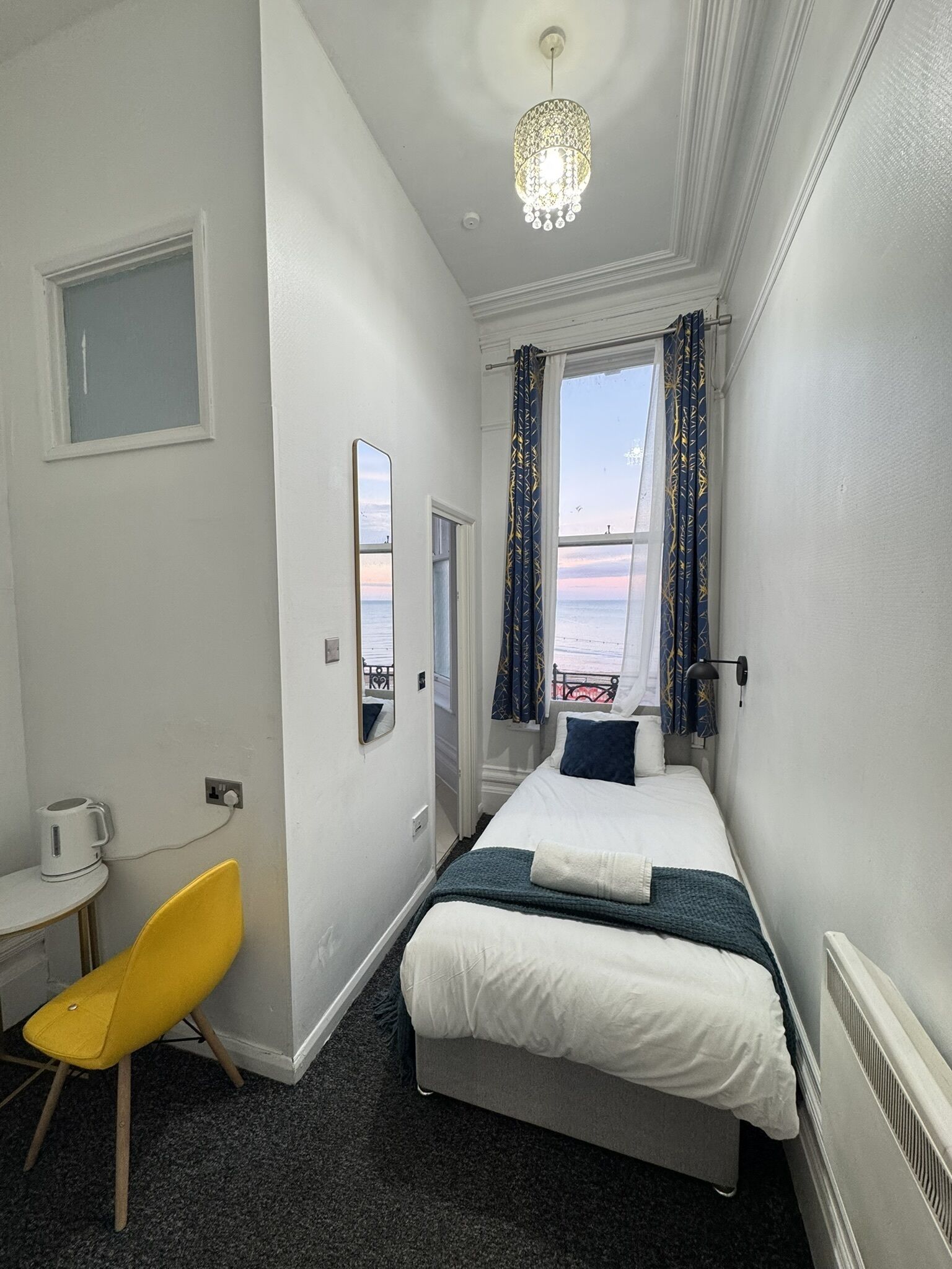 A hotel room with a bed, a chair, a small table, a mirror, a window with curtains, and a chandelier.