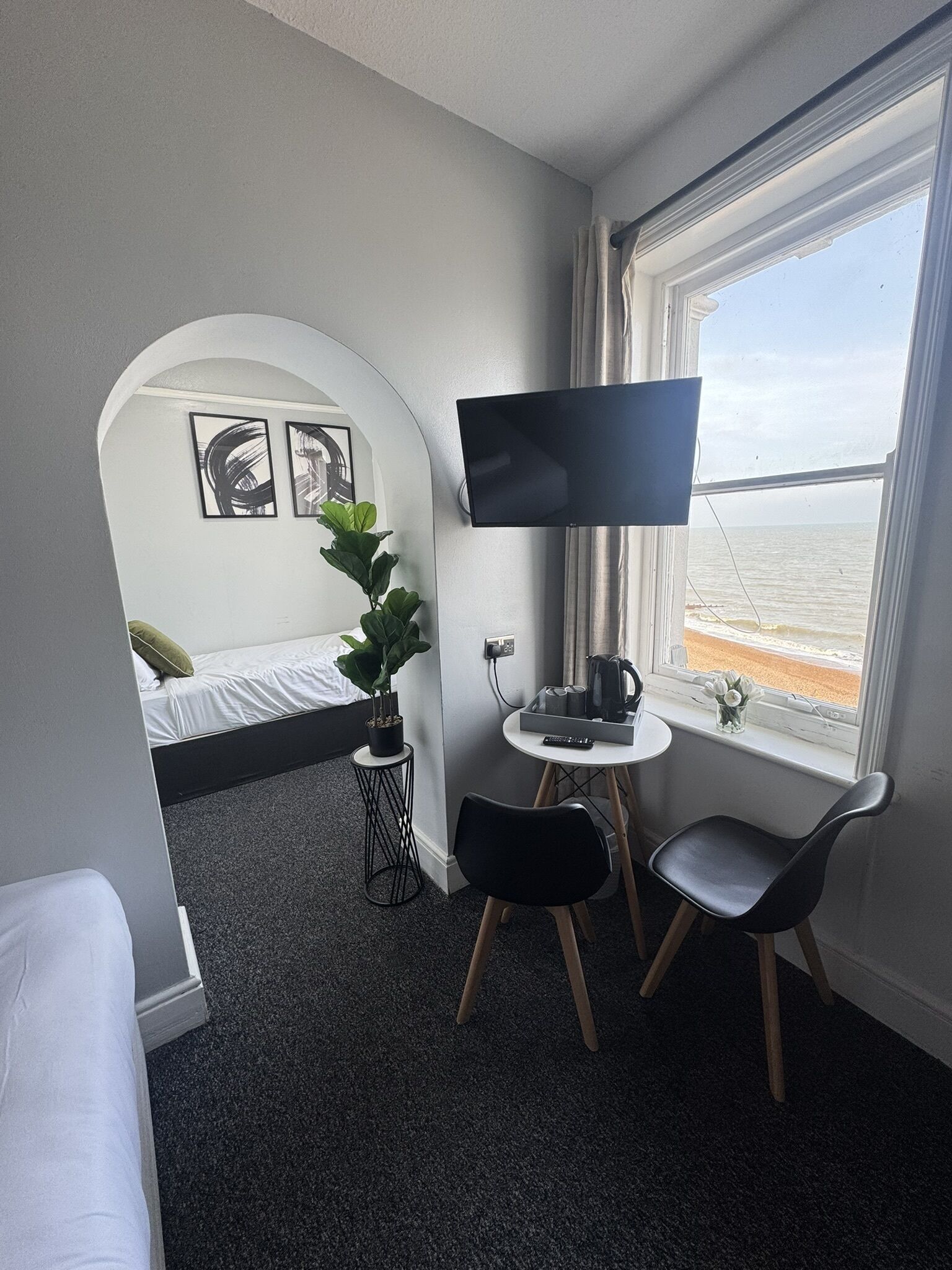 A compact hotel room with a bed, a desk with a TV, a small table, and a window with a view of the sea.