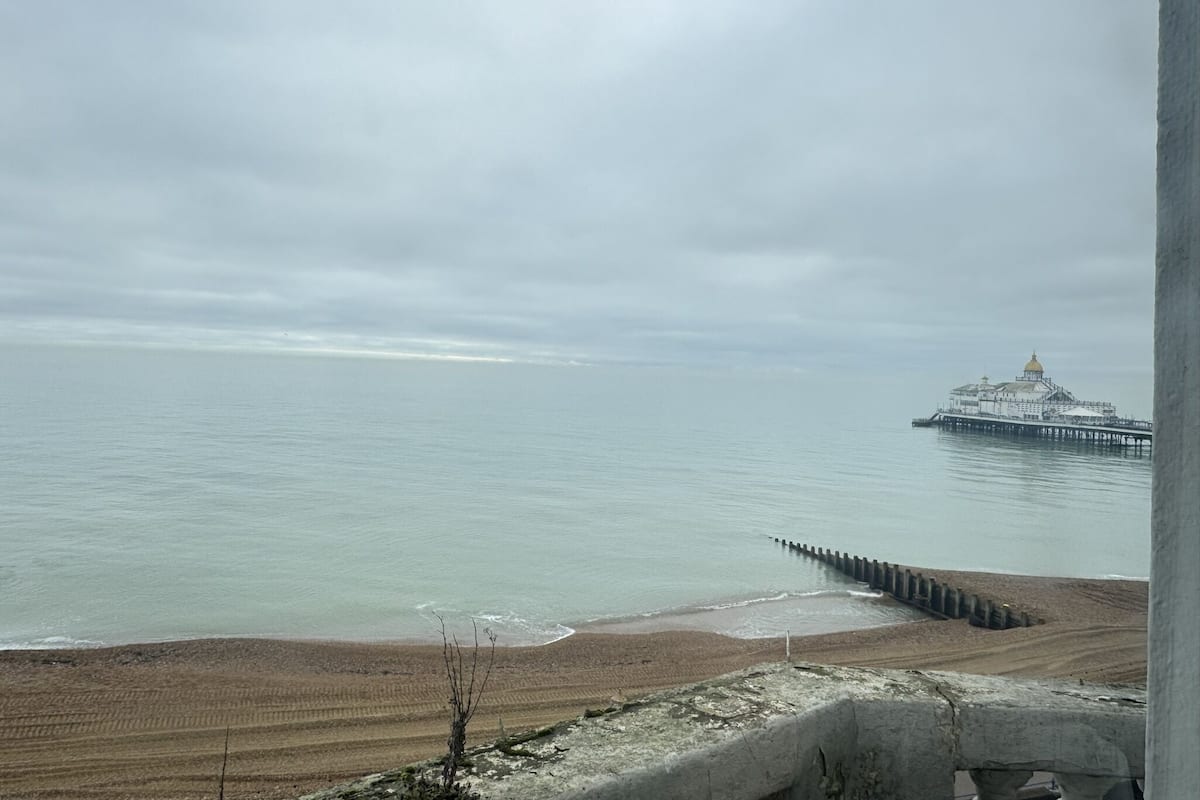 A coastal view with a pier extending into calm waters, a sandy beach, and a large white building with a golden dome.