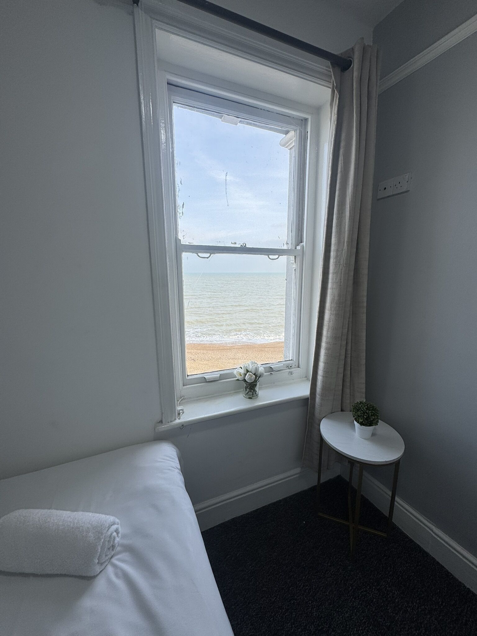 A room with a bed, a small table with a plant, a window with a view of the beach, and a curtain.