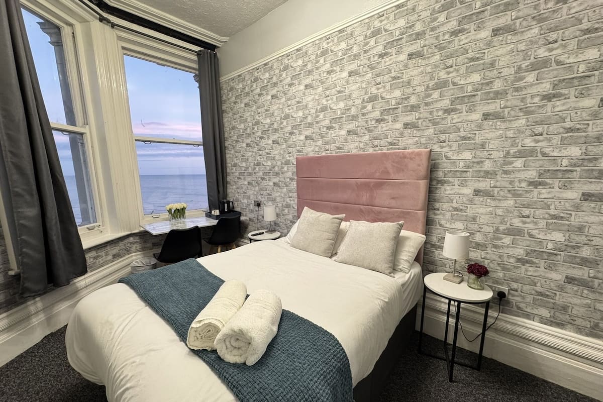A hotel room with a large bed, a view of the sea, a brick wall, and a small desk with a chair.