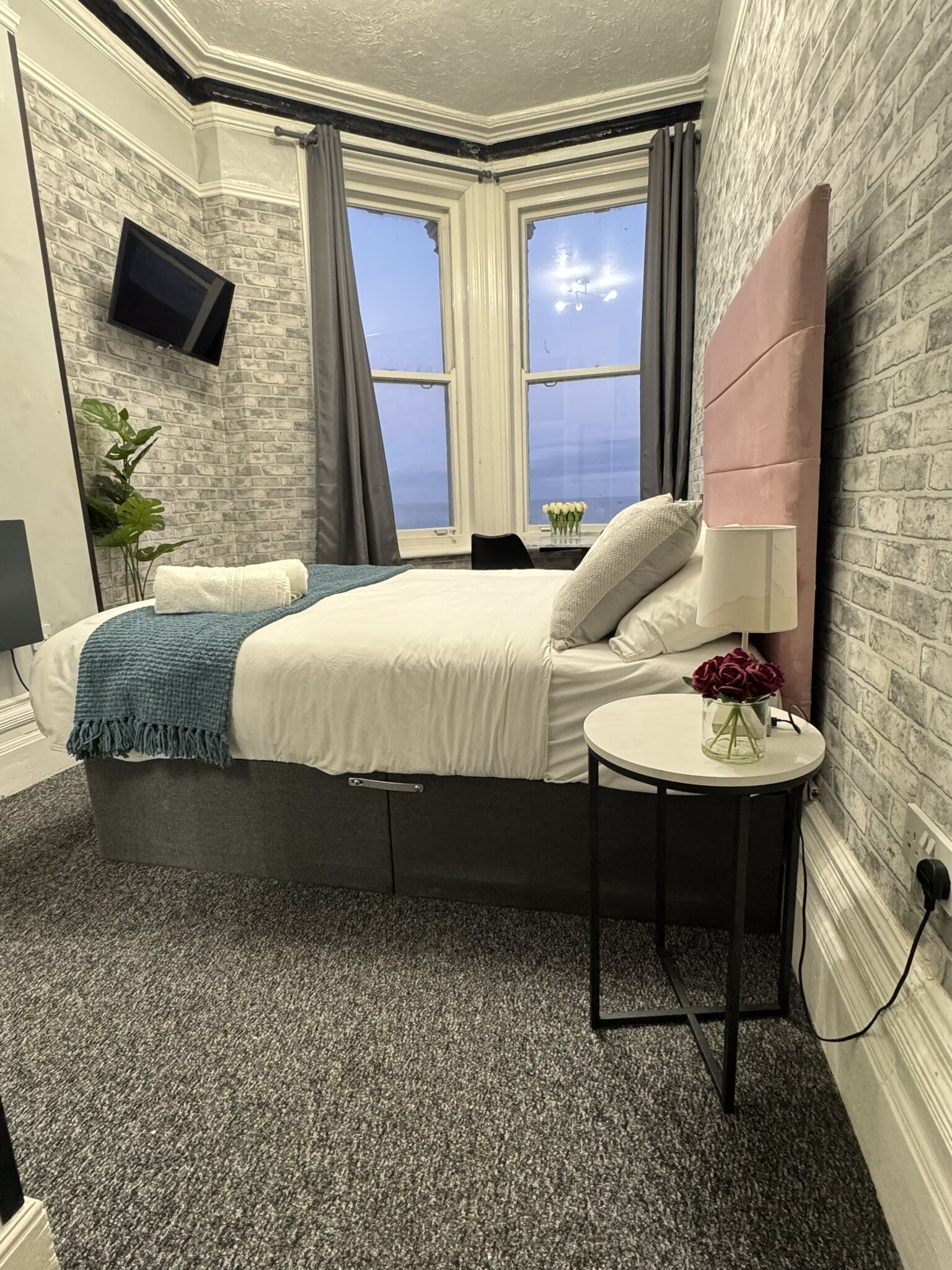 A modern bedroom with a large bed, a small table with a vase of flowers, and a view of the sky through a window.