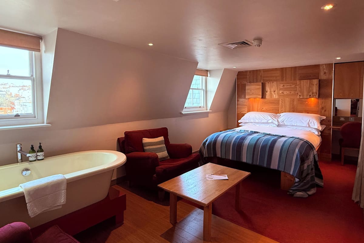 A bedroom with a large bed, a wooden headboard, a red armchair, a wooden table, and a bathtub.