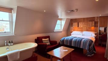 A bedroom with a large bed, a wooden headboard, a red armchair, a wooden table, and a bathtub.